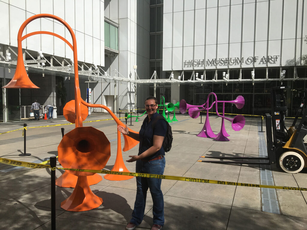 Liz stands within caution tape and gestures toward a bright orange many-horned sound sculpture.