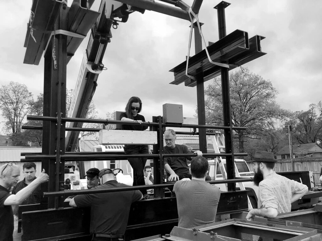 People stand around as a crane holds a catwalk component by a segment of I-beam.