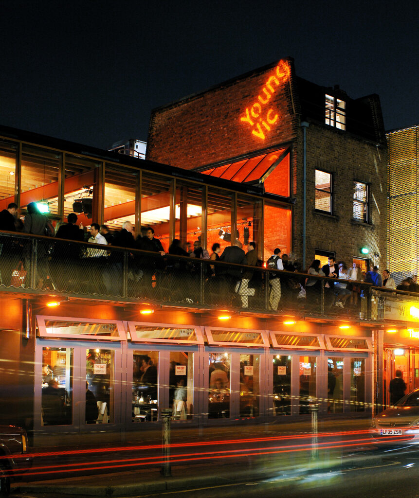 A long exposure photograph of Young Vic. People fill the area beyond the glass doors on the first floor and line the second floor balcony. The words Young Vic are projected onto the side of an old brick building.