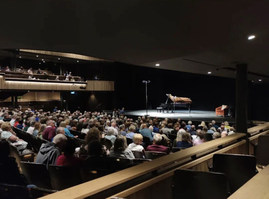 An audience watches a performance with a piano and lounge chair onstage.