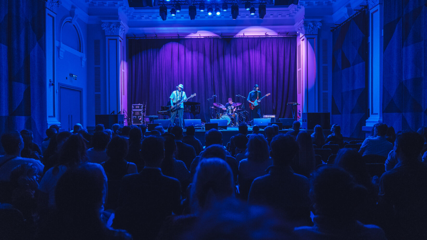 A band with guitar, bassist and drums performs on stage under blue lighting. An audience fills the room.