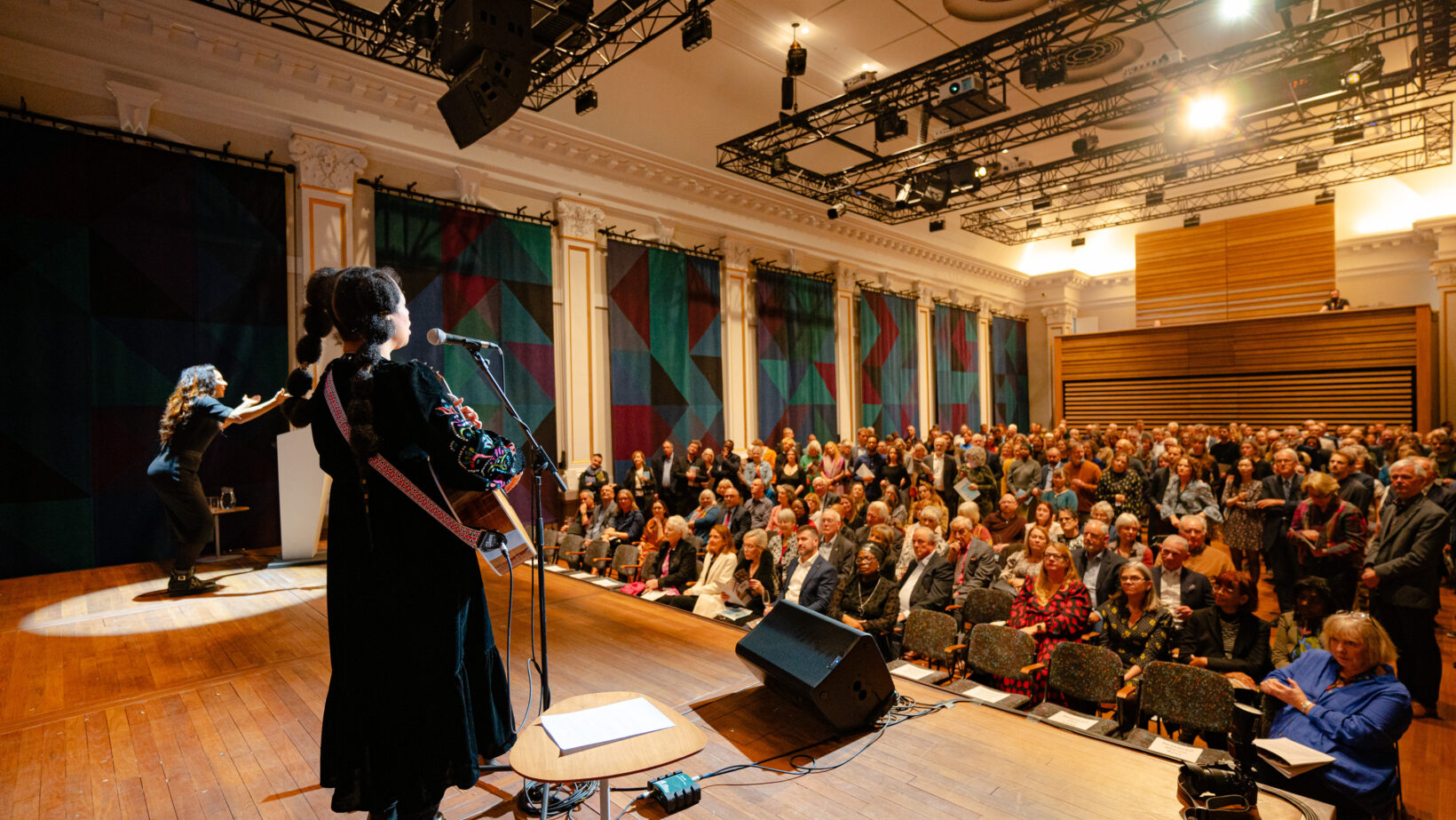 A performer in a black embroidered dress plays guitar before a full audience. Colorful tapestries hang along the wall.