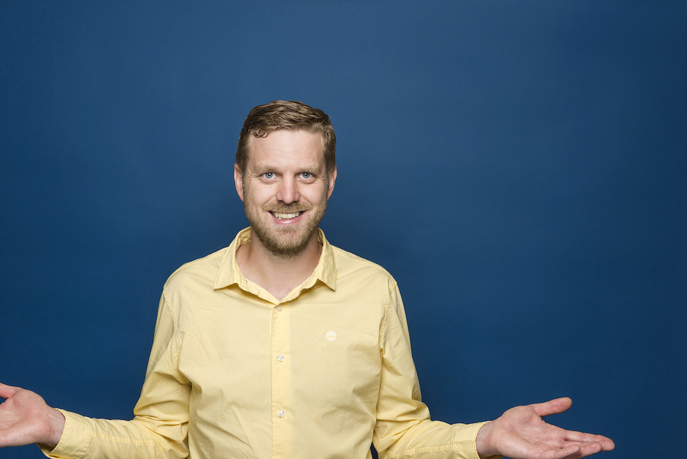 Ian Stickland in a yellow shirt, smiling, with arms at his sides outstretched.