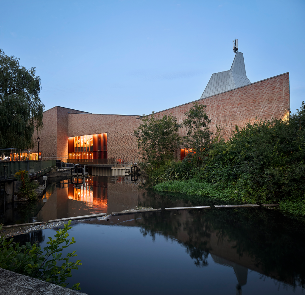 An angular brick building with illuminated window slats reflects in dark water.