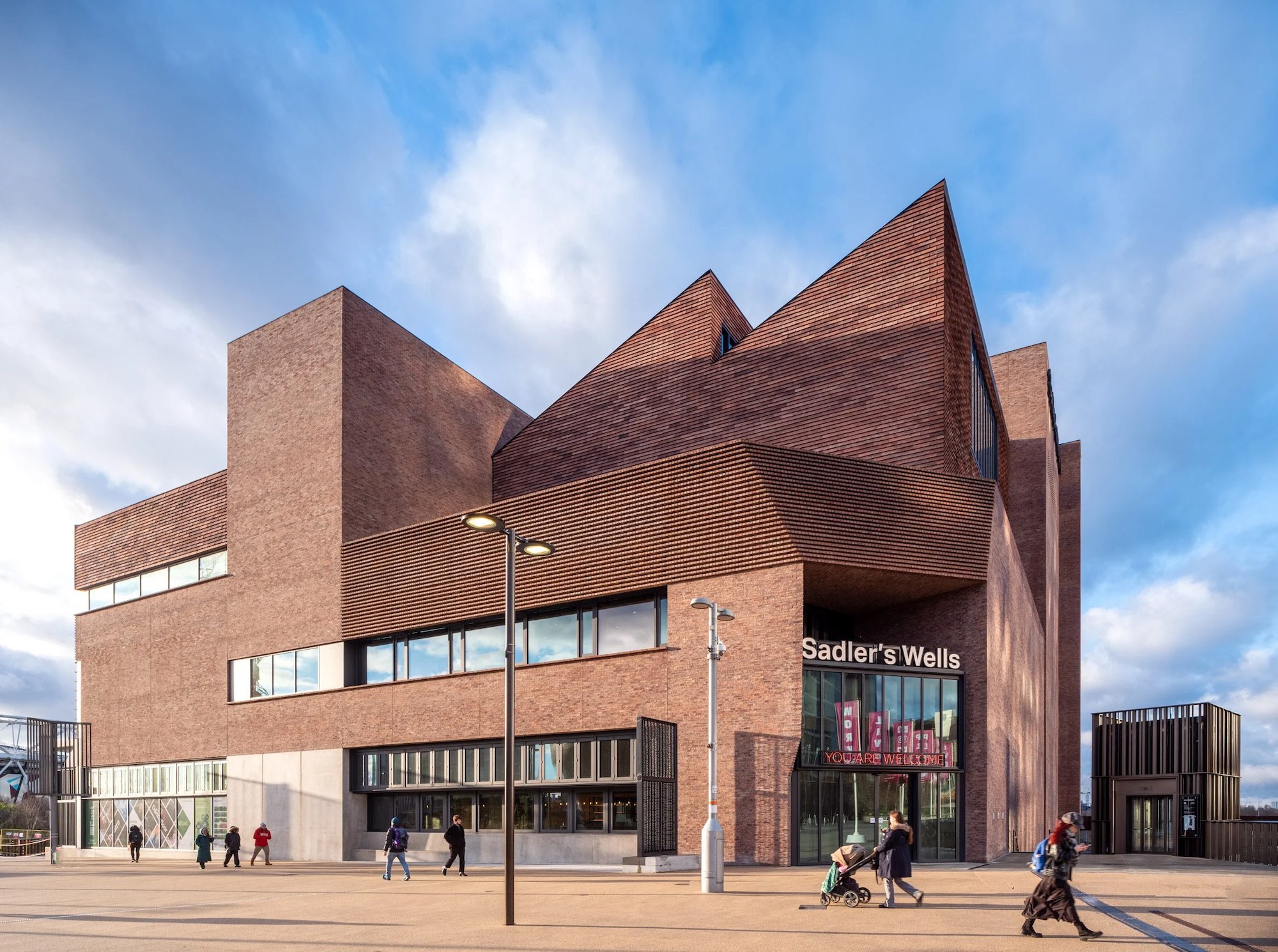 The brick exterior Sadler's Wells with jutting triangular roof. Pedestrians walk past. A neon sign says "You Are Welcome".