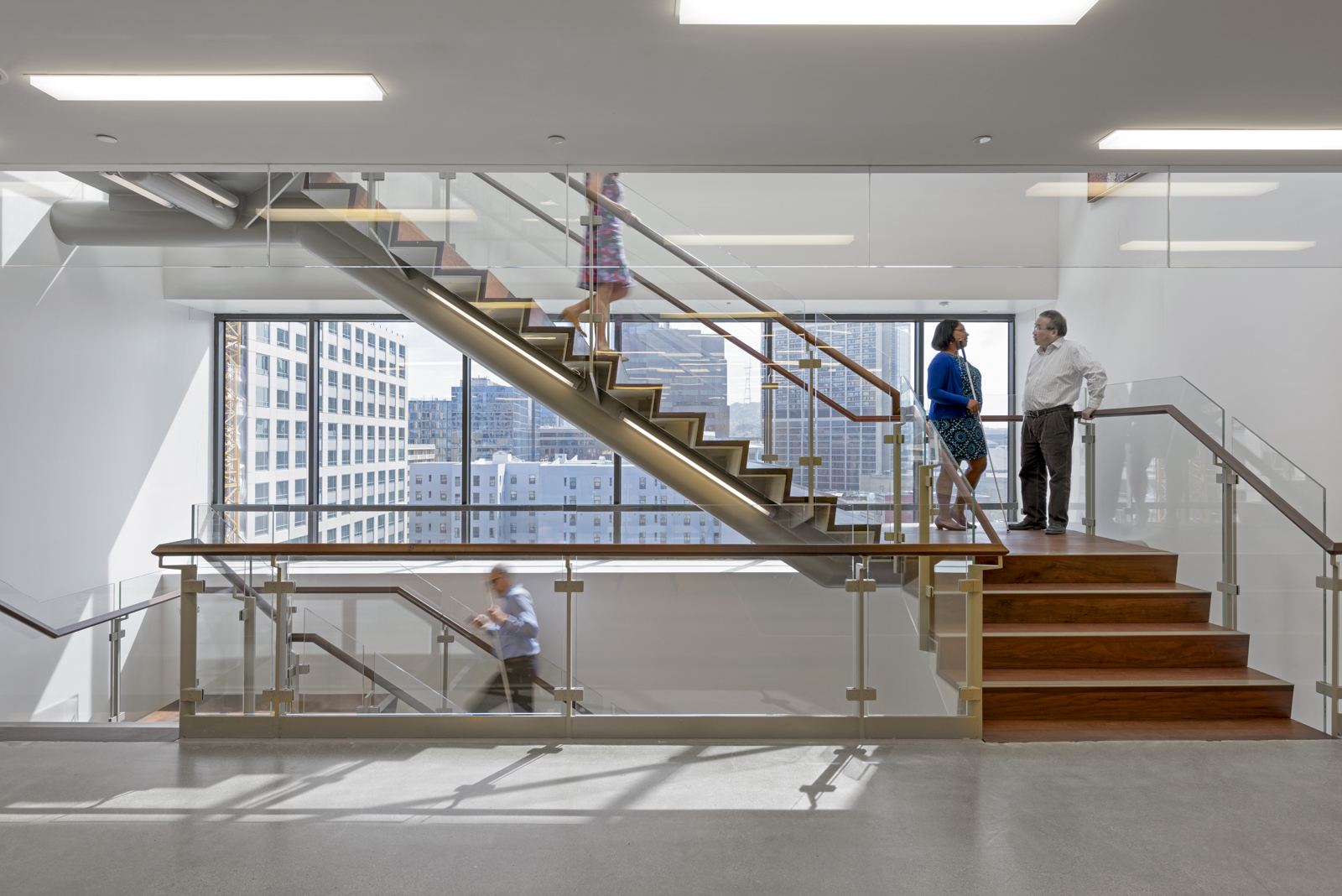 People climb and pause on the landings of a glass stairwell with continuous wooden railings.