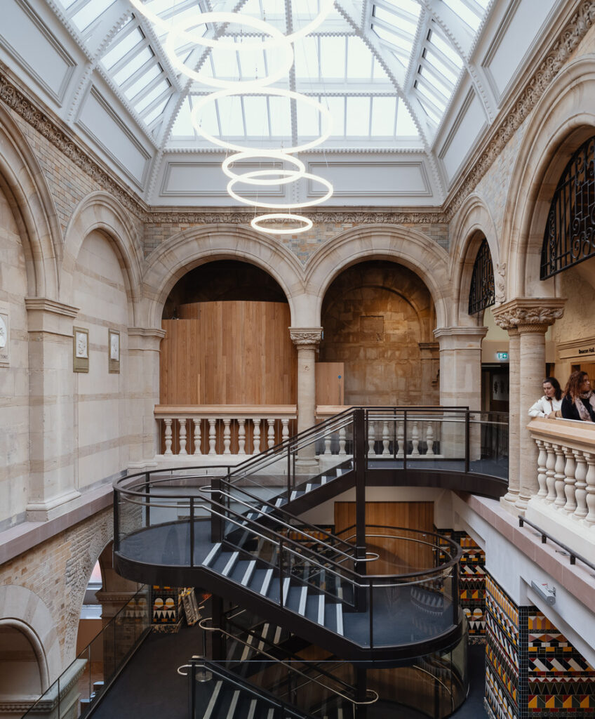 A contemporary metal and glass stairwell inside a historical looking foyer with columns, balcony, arches and decorative tile on the first floor.