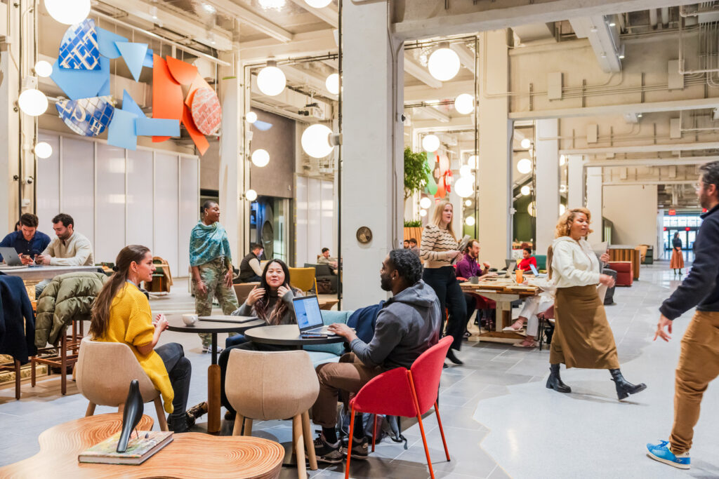 People gather with laptops and coffee in high-ceilinged collaborative space. Globular lights are scattered about the columns.