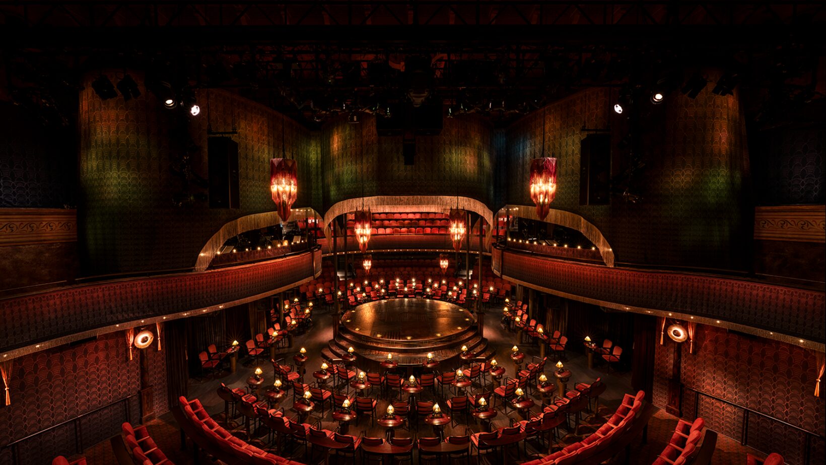 Chairs with tables and lamps surround a circular stage. The room is lit with dim red chandeliers.