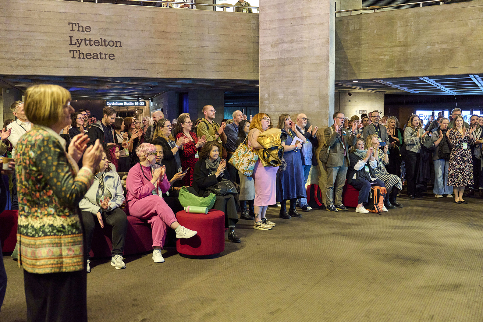A crowd on the ground floor an industrial space claps and looks on. A sign on the left reads The Lyttelton Theatre.