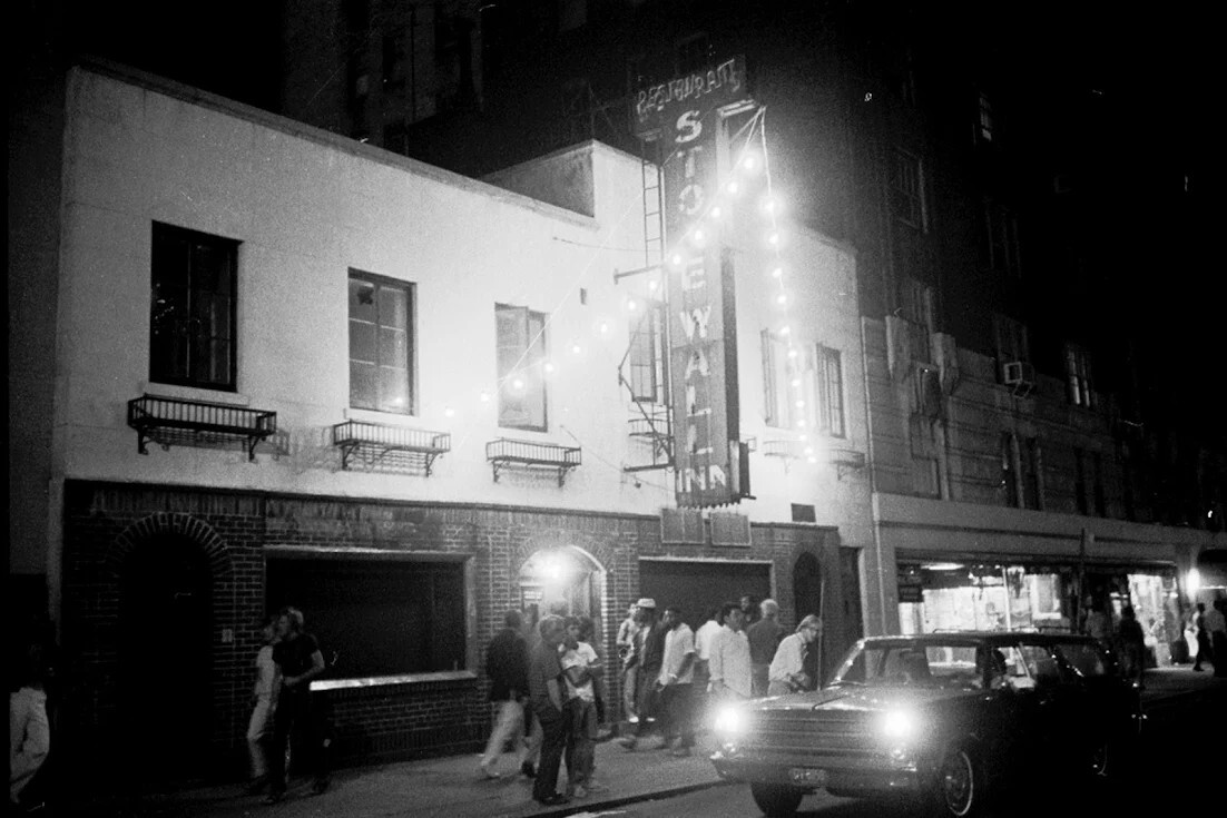 Black and white photograph of Stonewall Inn. People stand outside under the blade sign. The windows are blacked out.