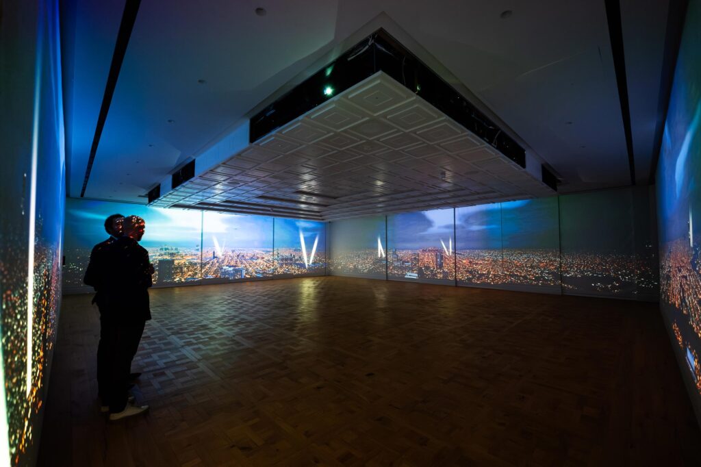 Two people stand in a room. A system of projectors protrude as a block from the ceiling and illuminate the walls with imagery of a city seen from above.