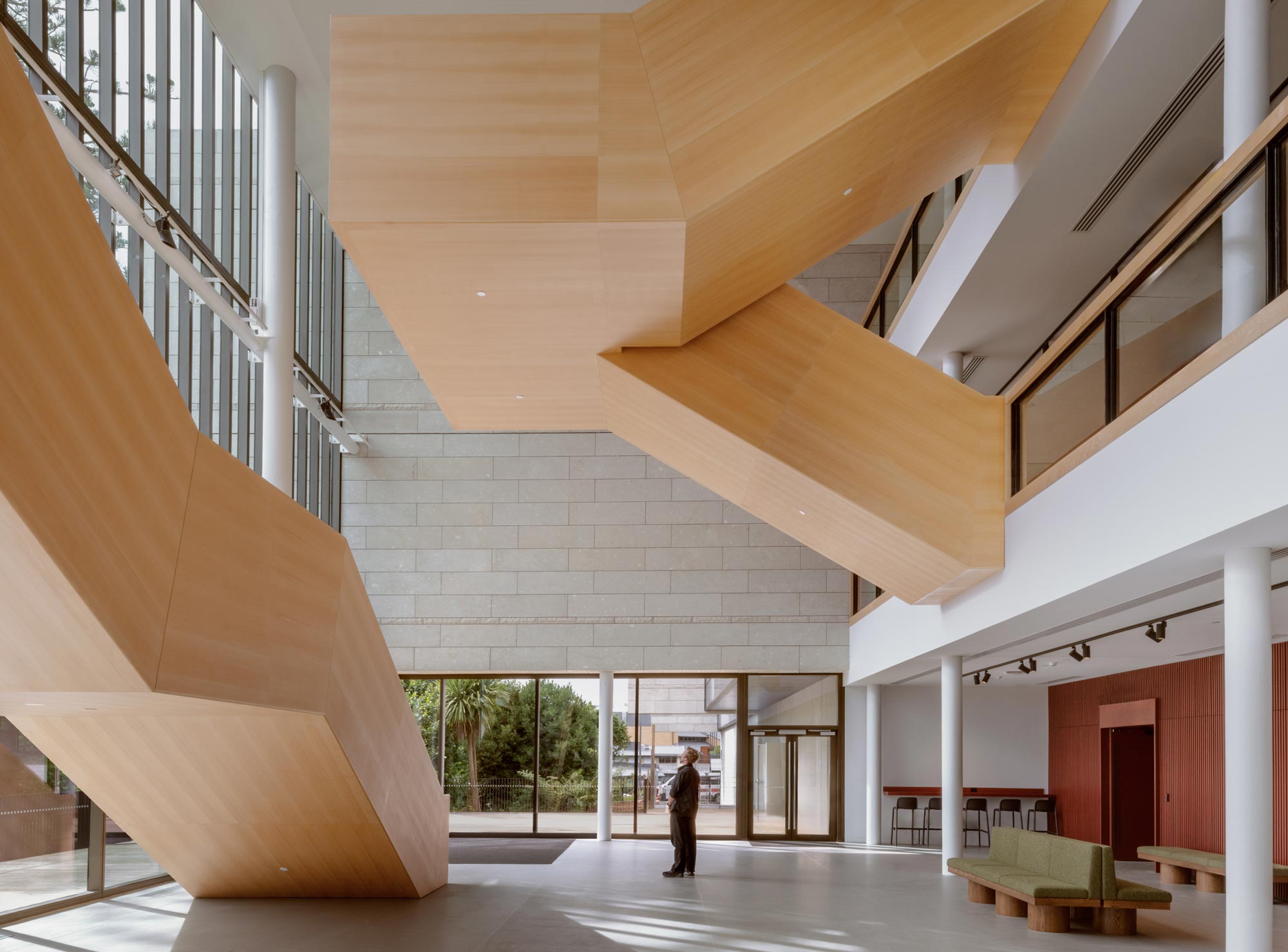 A person standing in the lobby looks up at the blocky wood veneer stairwells. Photography courtesy of Jasmax.