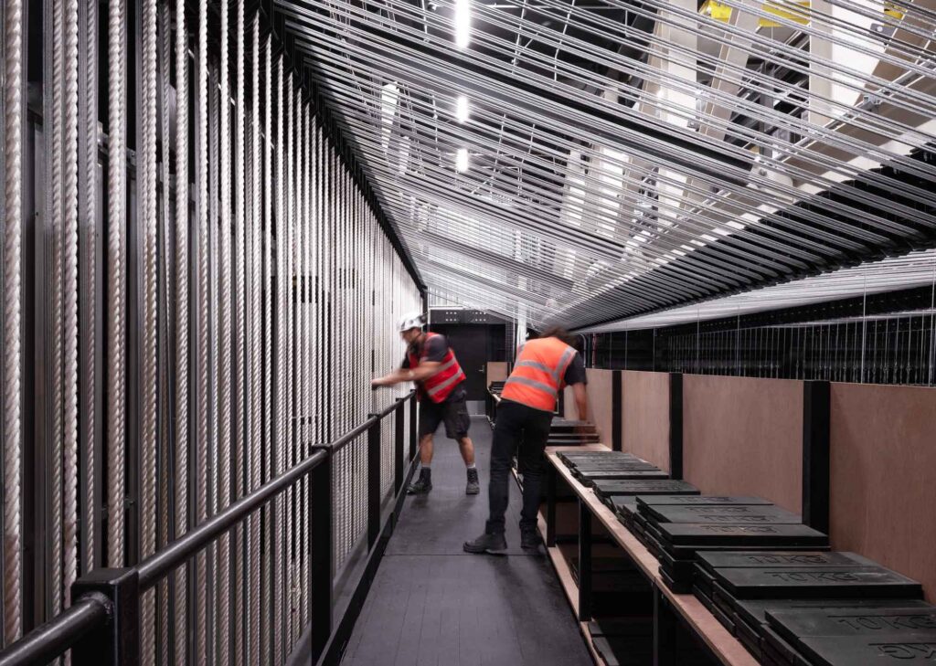 From a catwalk, people work with a massive wall of ropes and 10 KG weights.