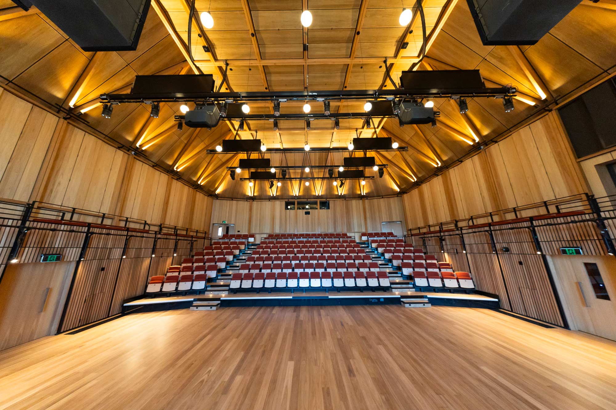 Raised seating in a wooden paneled room. The ceiling is lined with rows of speakers.
