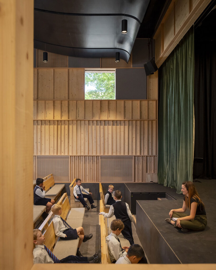 A half dozen young boys sit and stand on padded wooded seats. A woman sits cross-legged on the stage. A green curtain hangs to one side. An acoustic panel curves down toward the stage. The rear wall has natural wood finish with names engraved among the wooden slats.