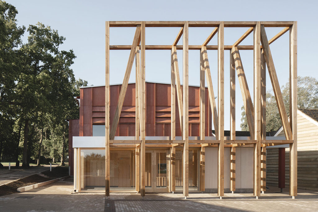 A tall wooden frame looms over the brick-red and glass theatre building, creating a congregating area outside the entrance.