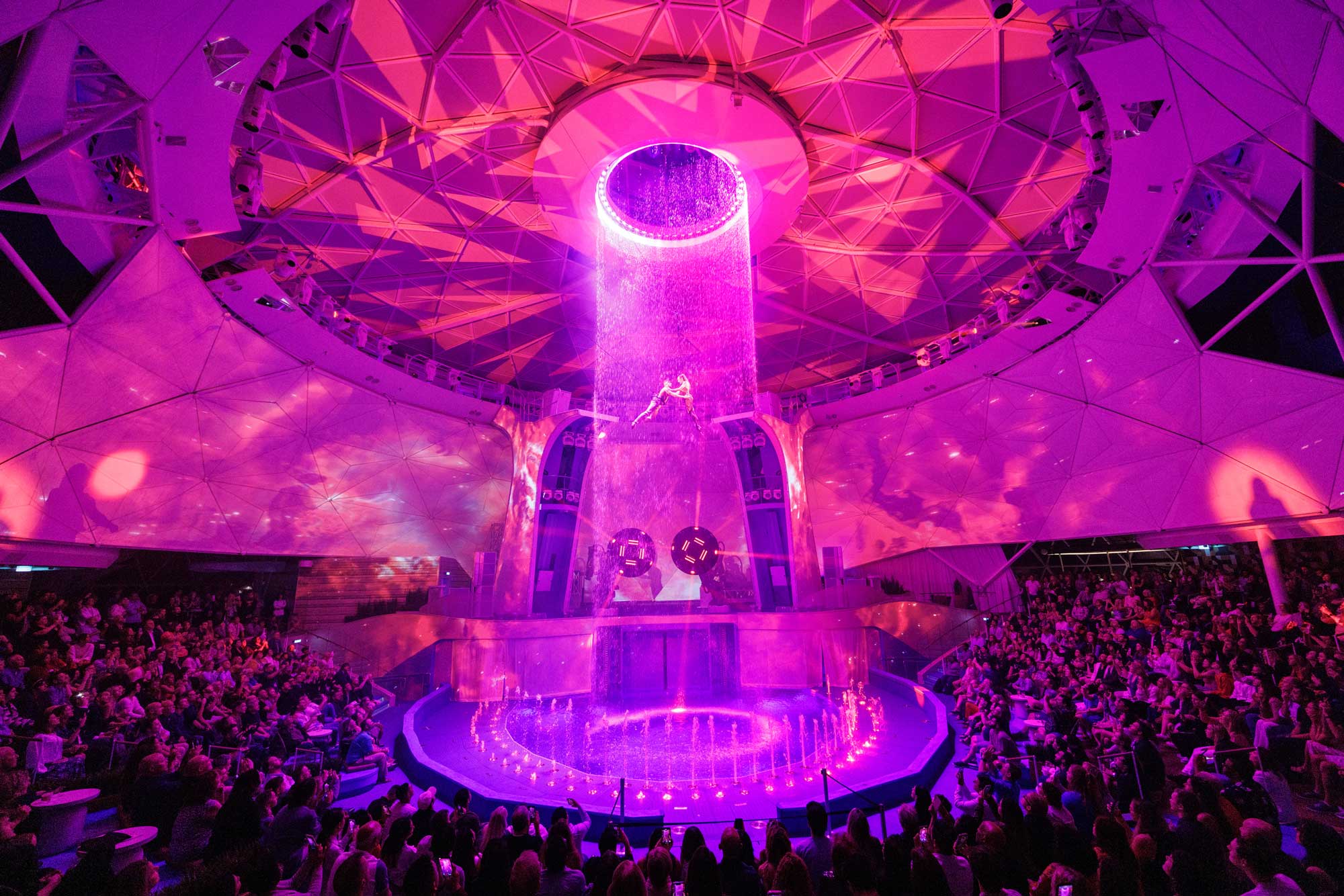 Water falls from a ceiling feature in the aquadome while performers hang suspended.