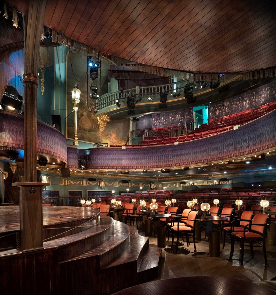 Pairs of chairs with individual tables and lamps surround a circular stage with stairs leading up to it. Rows of balconies are hung with golden fringe and moulded elements.