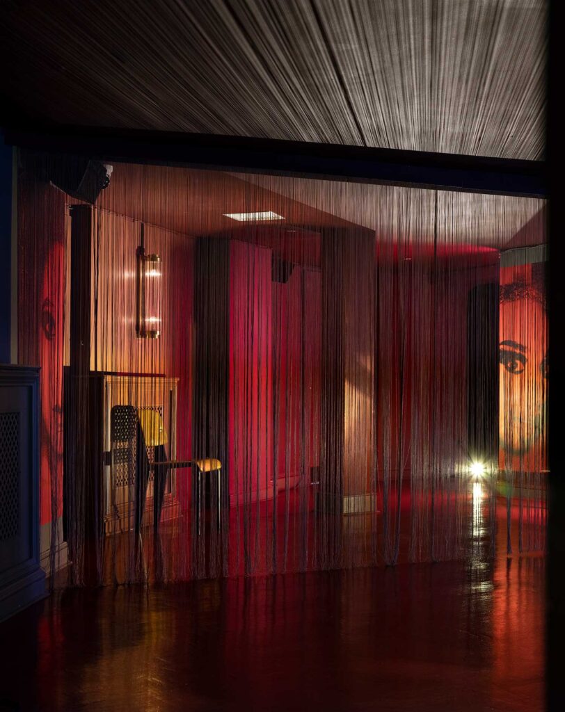 A curtain of threads casts a dramatic shadow over the ceiling. Floor to ceiling faces in dramatic makeup peer out from the walls of the red lit room.