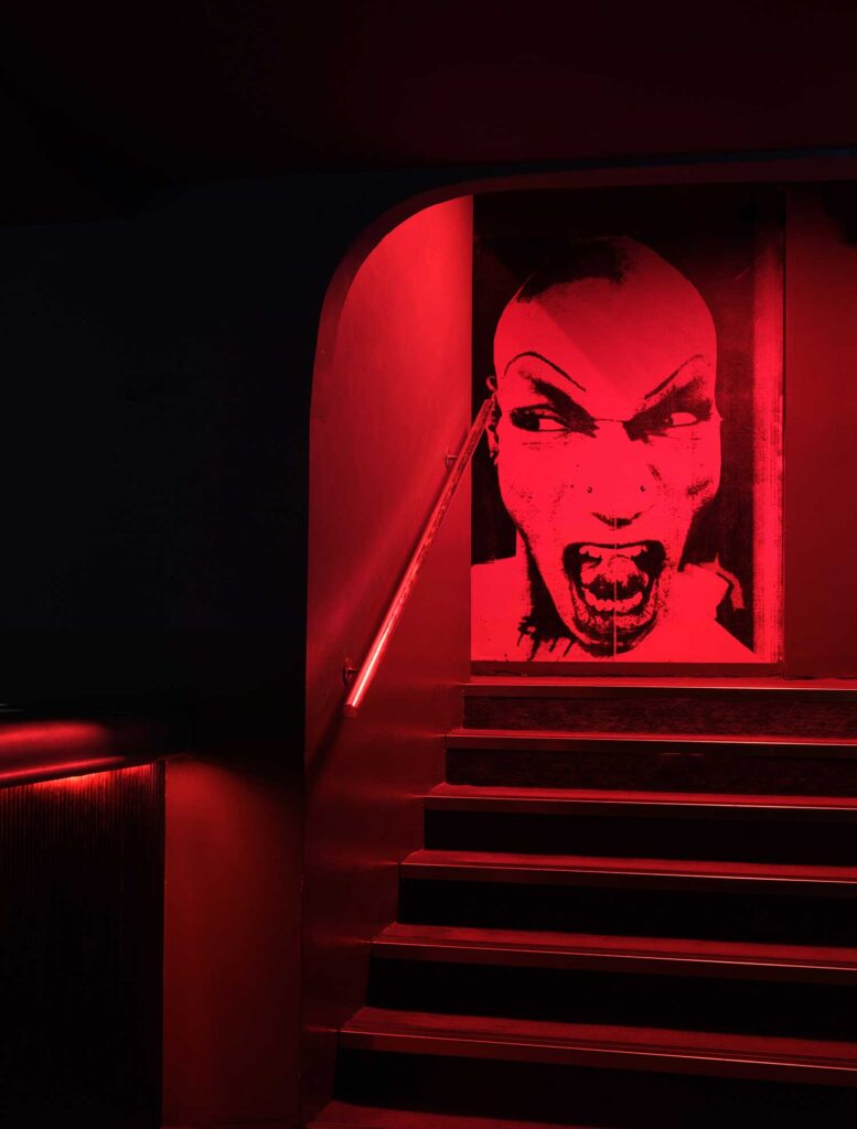 A red stairwell leads to a lurid image of a person with shaved head and tongue between teeth.