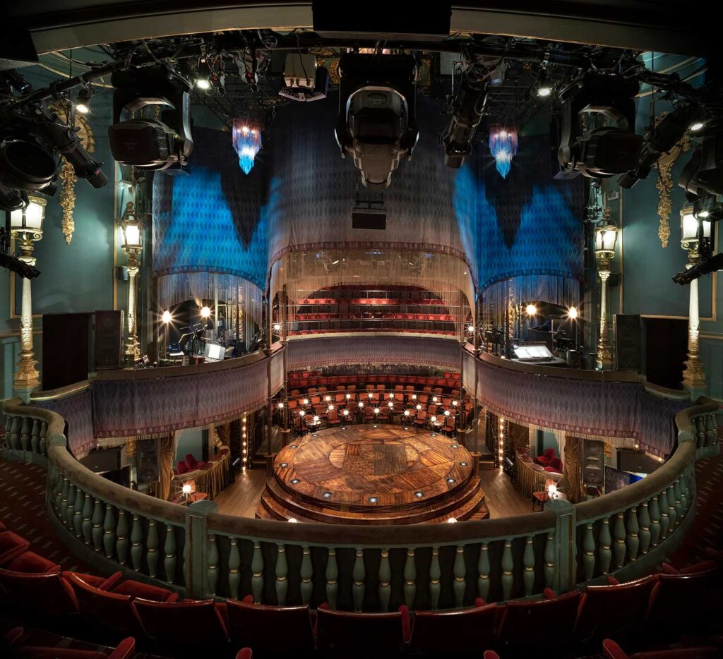 A view of the stage from the fringe-draped balcony level. Chandeliers are illuminated blue at the ceiling.