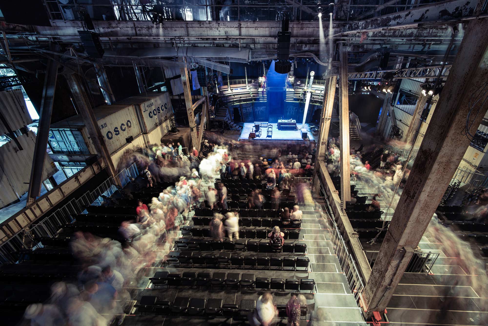 Long exposure image of audience filing through performance space amid COSCO shipping containers.