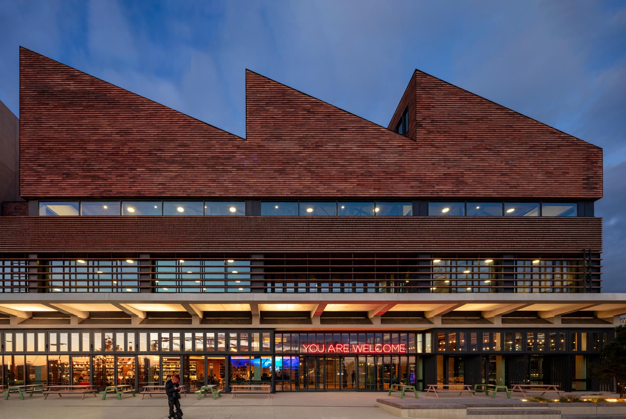 The brick exterior Sadler's Wells with jutting triangular roof. Pedestrians walk past. A neon sign says "You Are Welcome". © Nick Kane