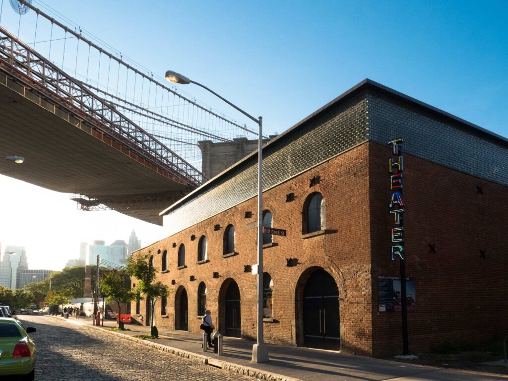 The brick exterior of St. Ann's Warehouse with arched doorways and stacked neon sign on the corner.