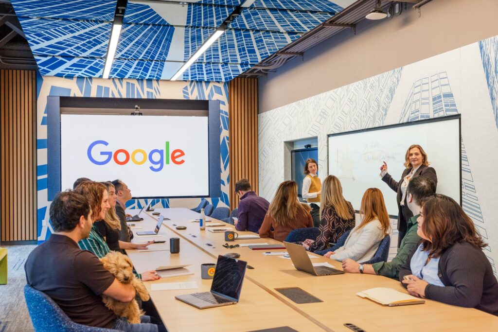 A team gathers in a meeting room with blue, urban wall and ceiling graphics, and digital screen on the wall.