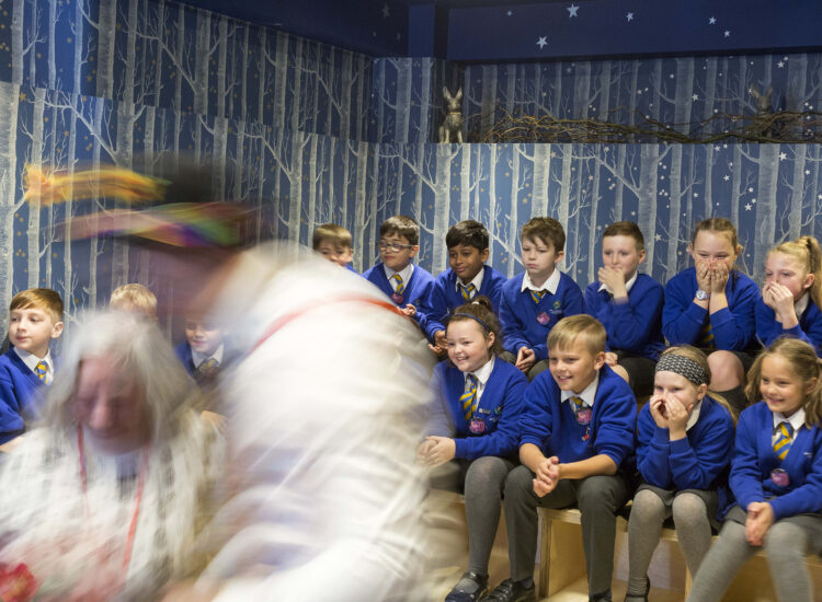 Children in blue uniforms smile and cover their faces as they watch a performance.
