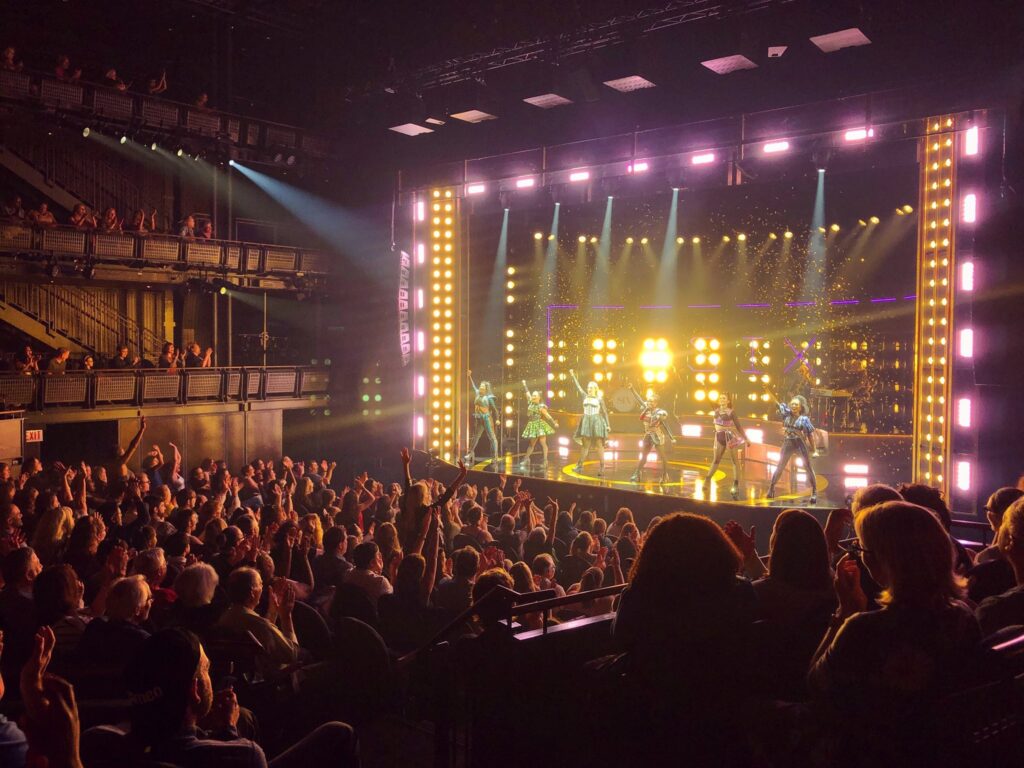 Performers pose with arms raised while illuminated by lights. The audience raises their arms and cheers.
