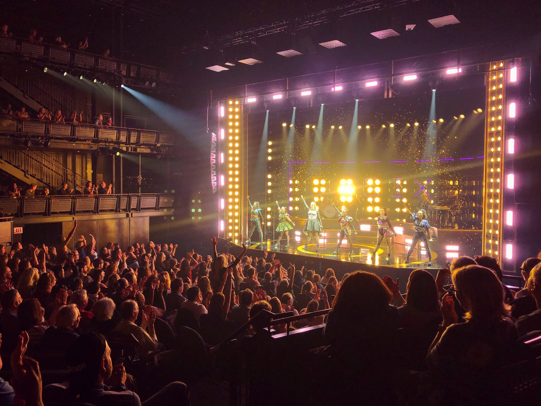 Performers pose with arms raised while illuminated by lights. The audience raises their arms and cheers.