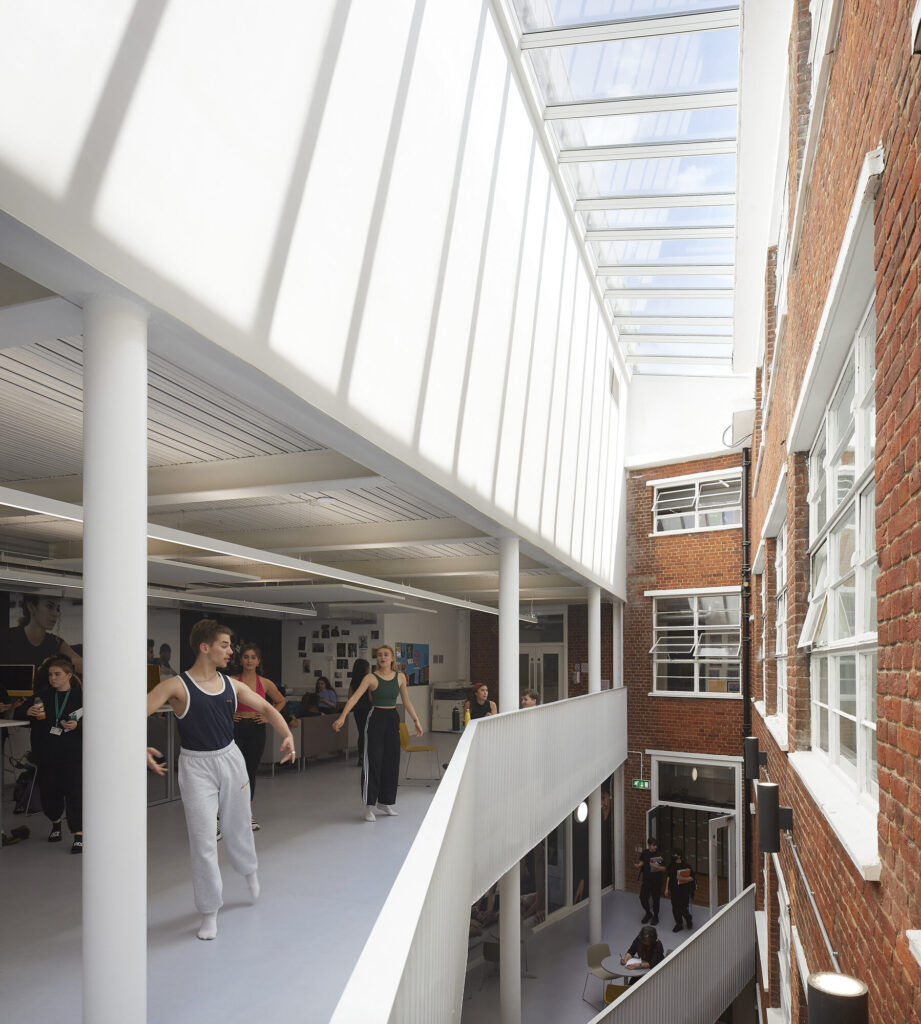 Dancers look down over a balcony toward rustic brick building facade. Image by Hufton & Crow.