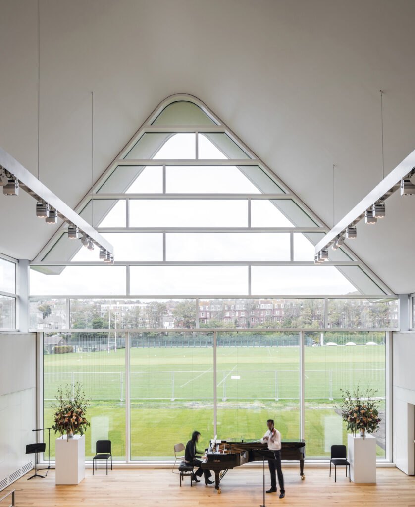 Students play piano and violin in front of expansive two-story window.