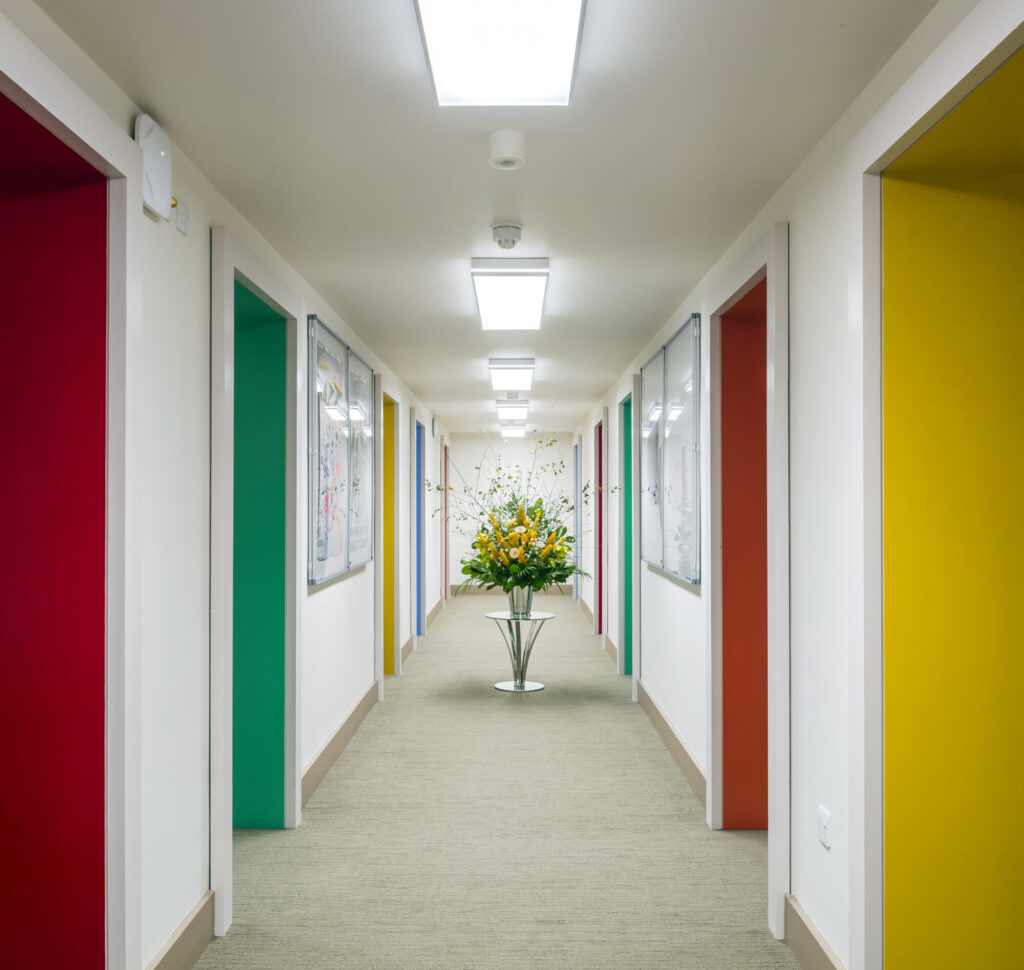 A white hallway with red yellow, green and blue doorways and a bouquet of flowers.