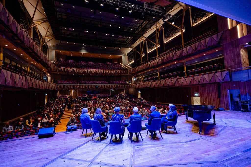 Seven people sit on stage under blue light before an audience at Bristol Beacon.