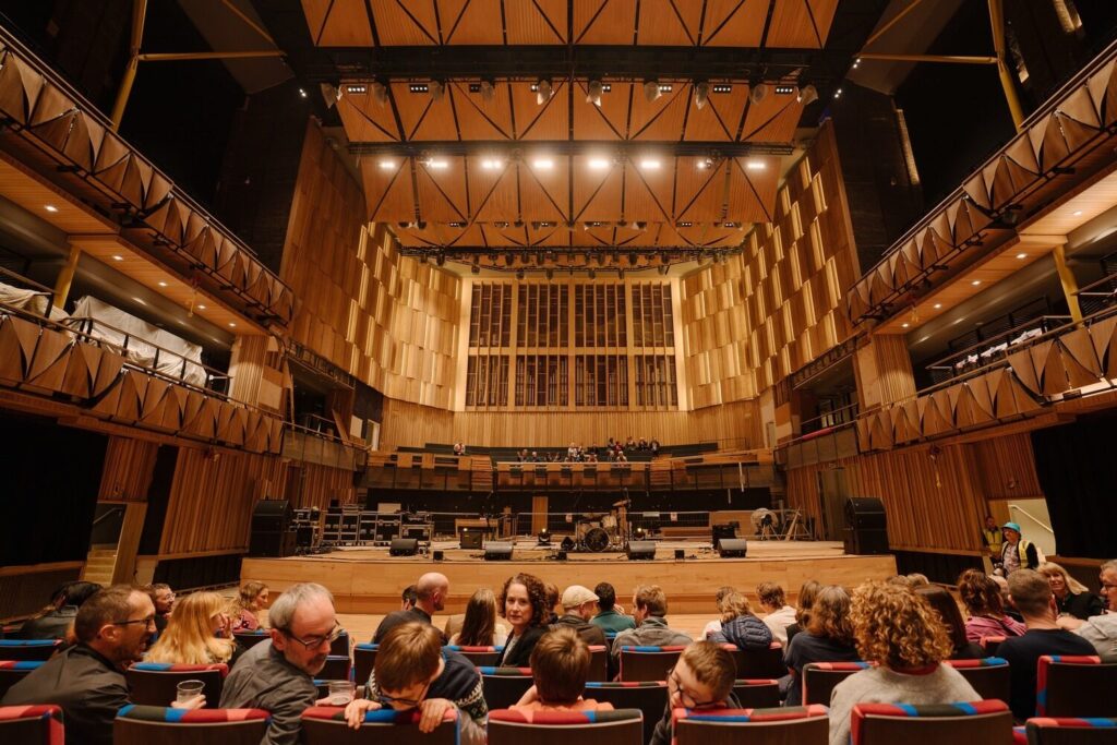 The audience takes their seats in the high-ceilinged performance space. Wood panels create an intricate raised pattern on the balconies and walls in the front.