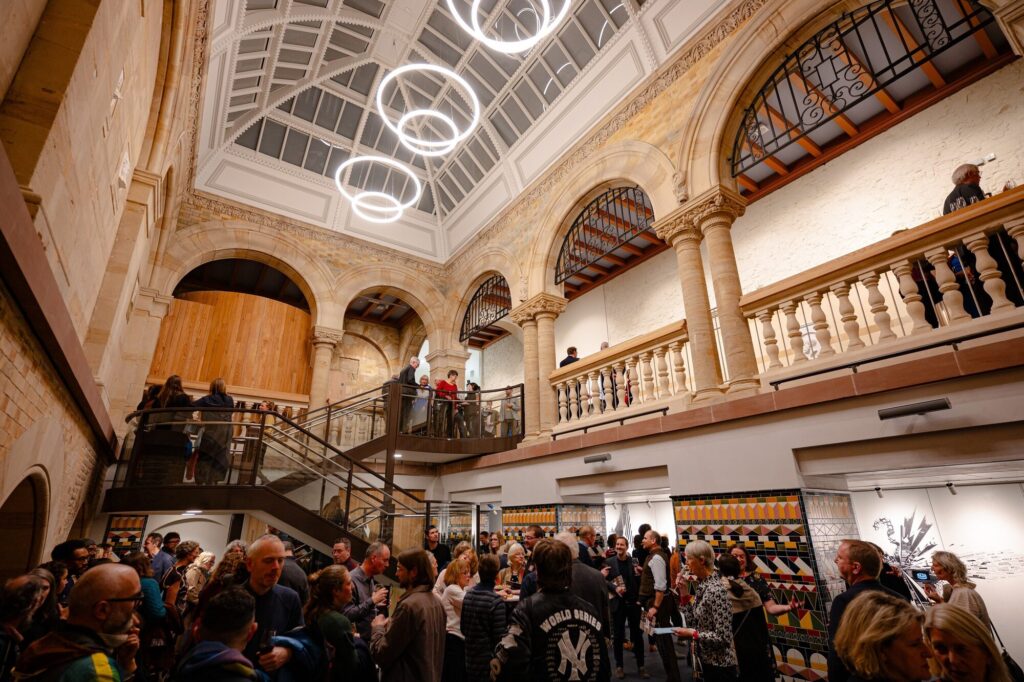 Visitors crowd the first floor, surrounded by elegant stone and tilework. Some visitors climb stairs to the second level. Contemporary circular lights hang from the ceiling.