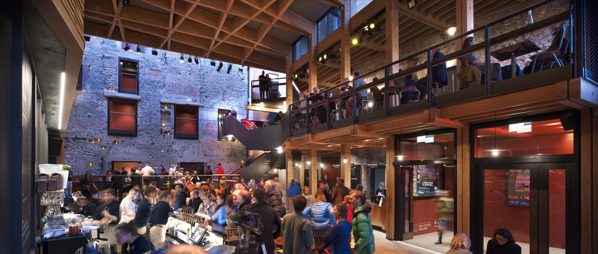The crowded bar and box office at Bristol Old Vic. A Rustic brick wall with blacked out windows forms the rear wall. Stairs lead up to a contemporary looking balcony.