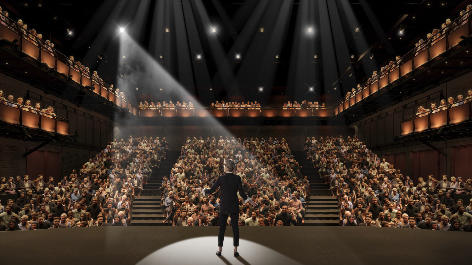 A person on stage before a crowded audience with two levels of balcony seating.