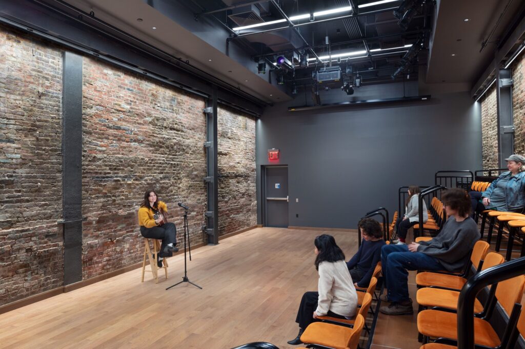 A person sits on a stool and speaks to audience in intimate performance space. Images by John Bartelstone Photography