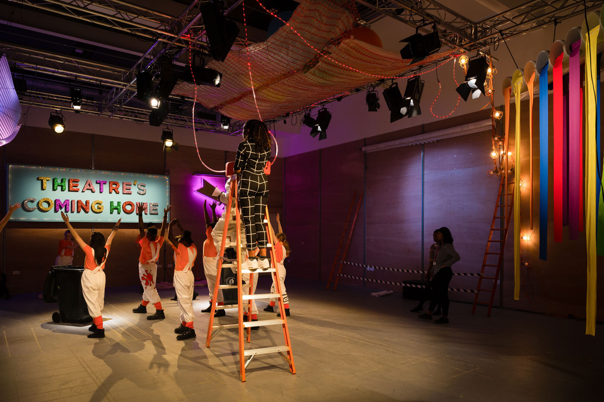 A woman on a ladder strings rope lights while performers raise their hands toward a sign that says 