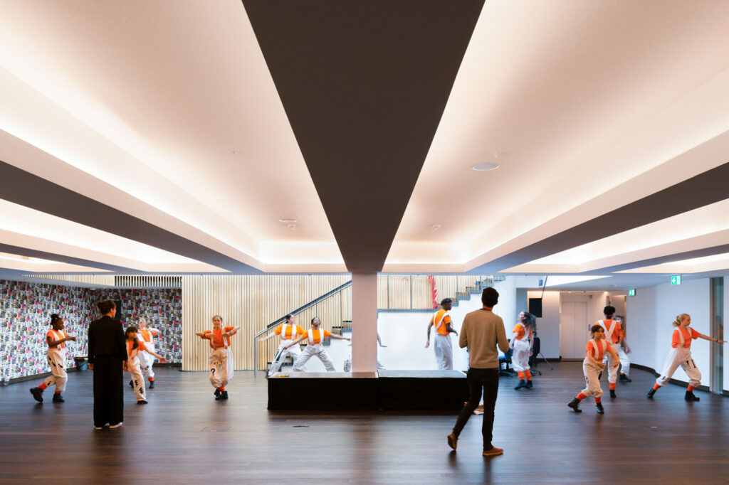 Young people in orange costume go through choreography in a room with stairwell.