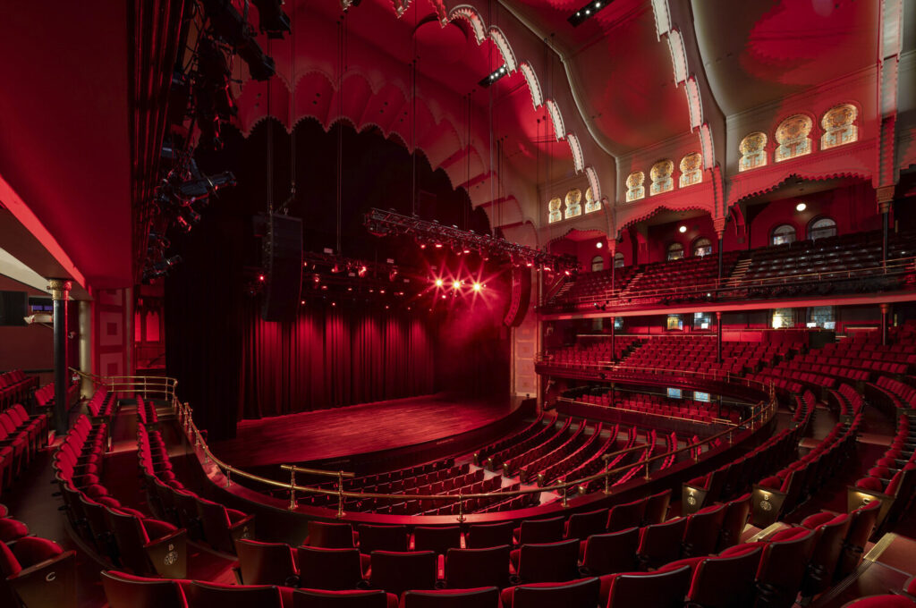 The red-illuminated interior of Massey Hall with elaborate ceiling features and stained glass windows. Photo by Scott Norsworthy.
