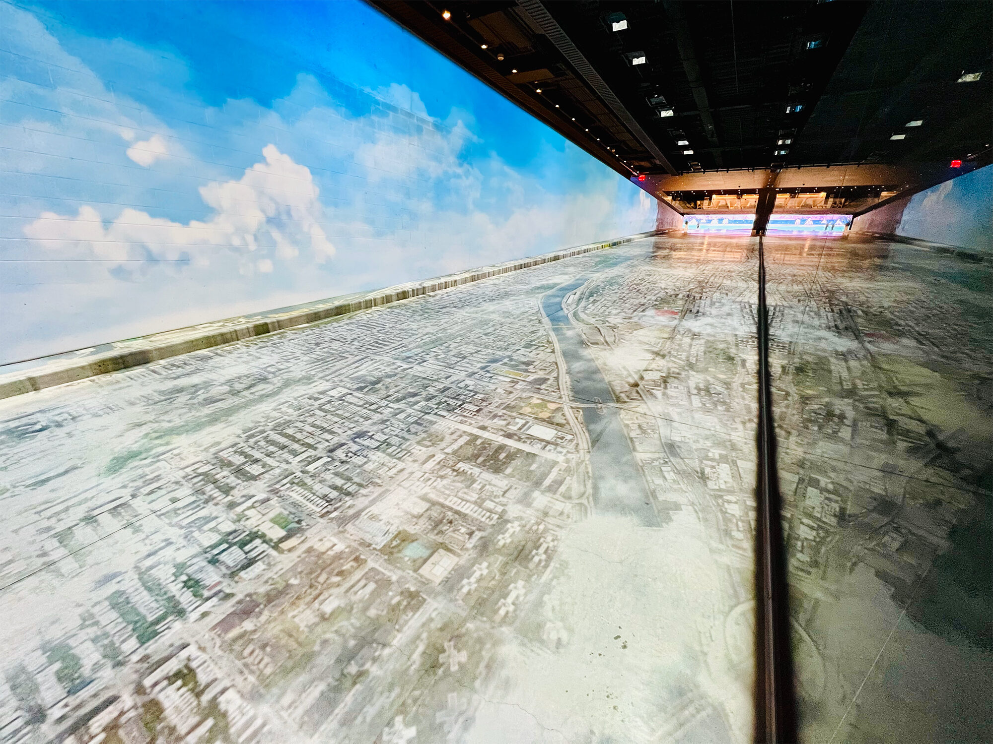 A long hall with a floor covered in aerial photography. The left wall is covered with artwork of a blue sky. The right wall is a mirror.