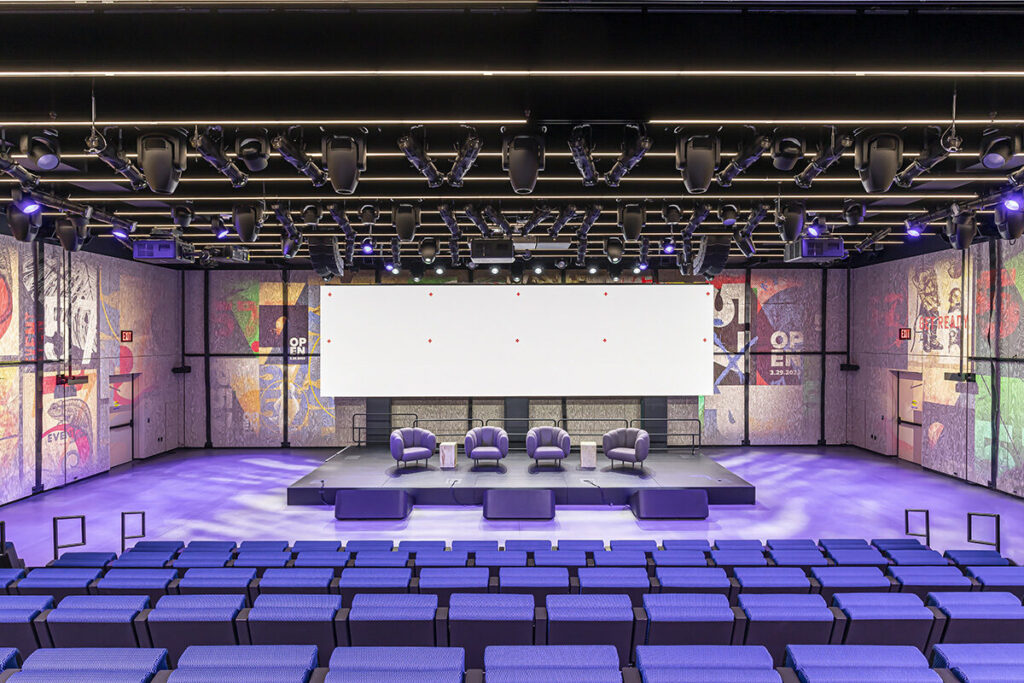 Four plush chairs on stage in front of purple bench seating. The ceiling is hung with several dense rows of AV equipment.