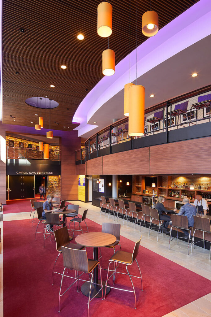 People sit at a bar in the lobby. A sign reads Carol Sawyer Stage at the rear of the space.