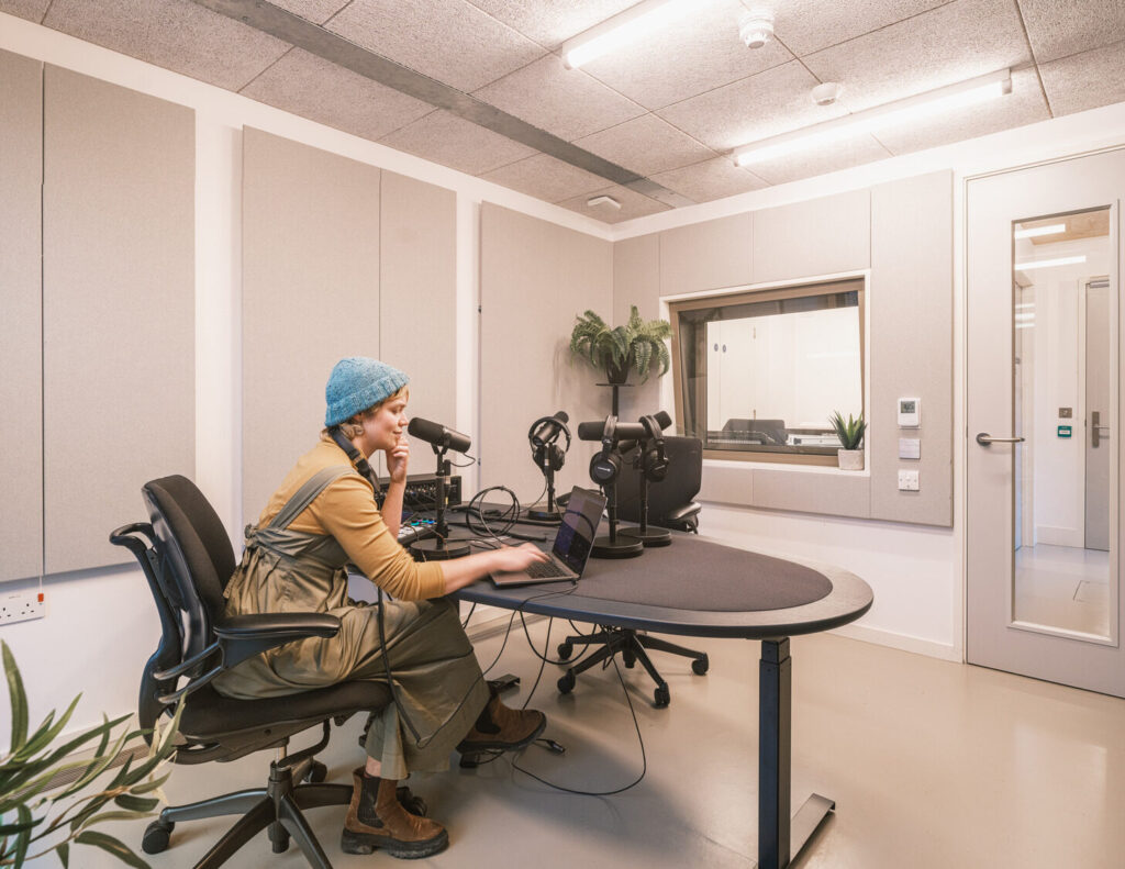 A person in a beanie works on a laptop in a private room. The table is stocked with sound recording equipment.