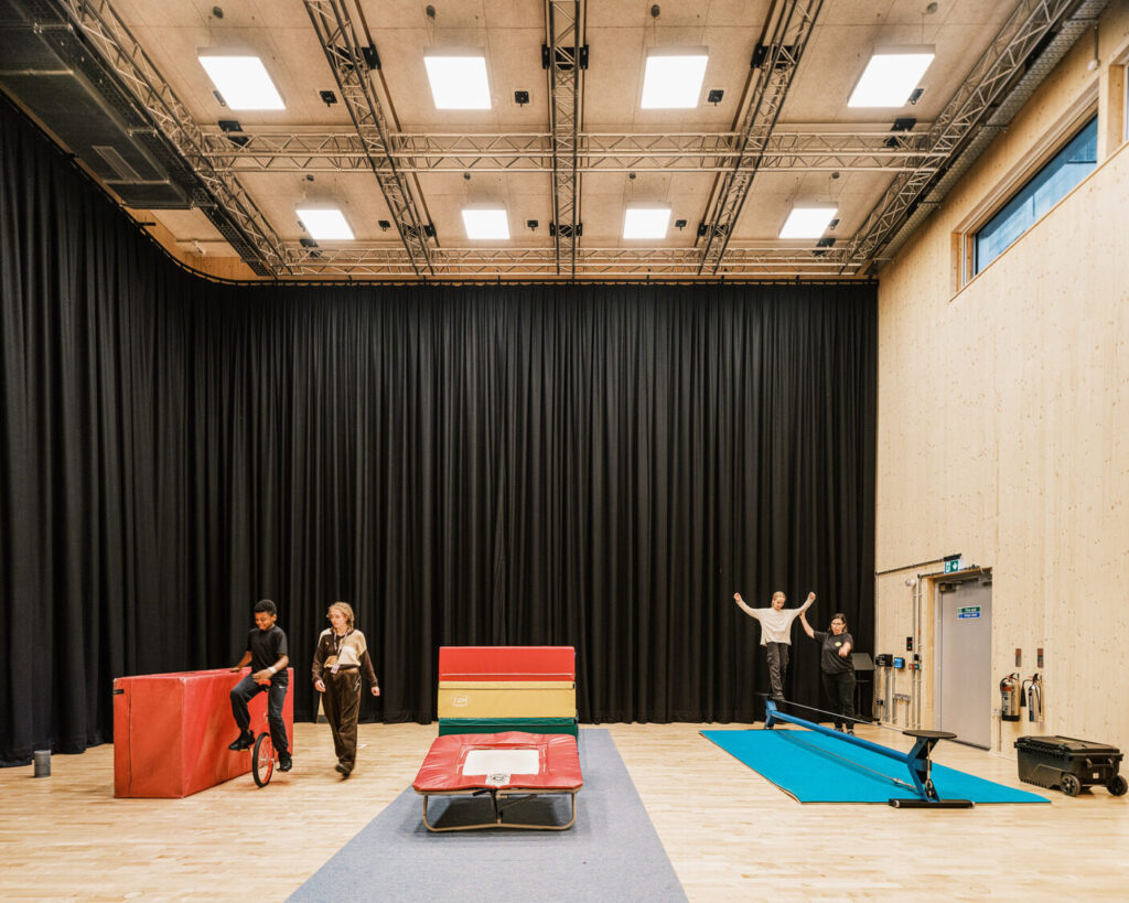 A boy rides a unicycle in a high-ceilinged room with a black curtain. Another student walks a tightrope.
